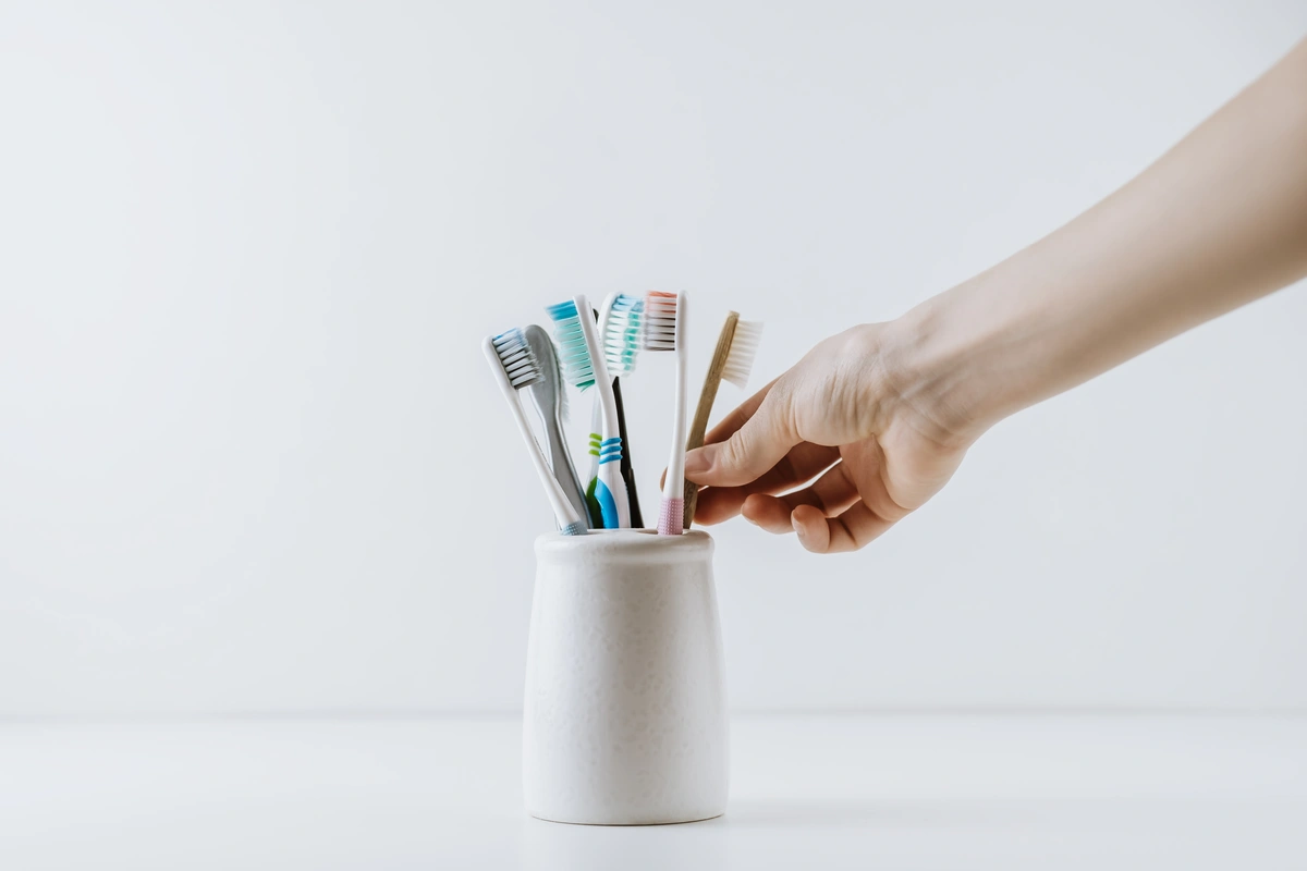 A hand selects an electric toothbrush from a white ceramic holder filled with various toothbrushes. The minimalist white background emphasizes the colorful heads, highlighting the choice between an electric toothbrush vs. a manual toothbrush.