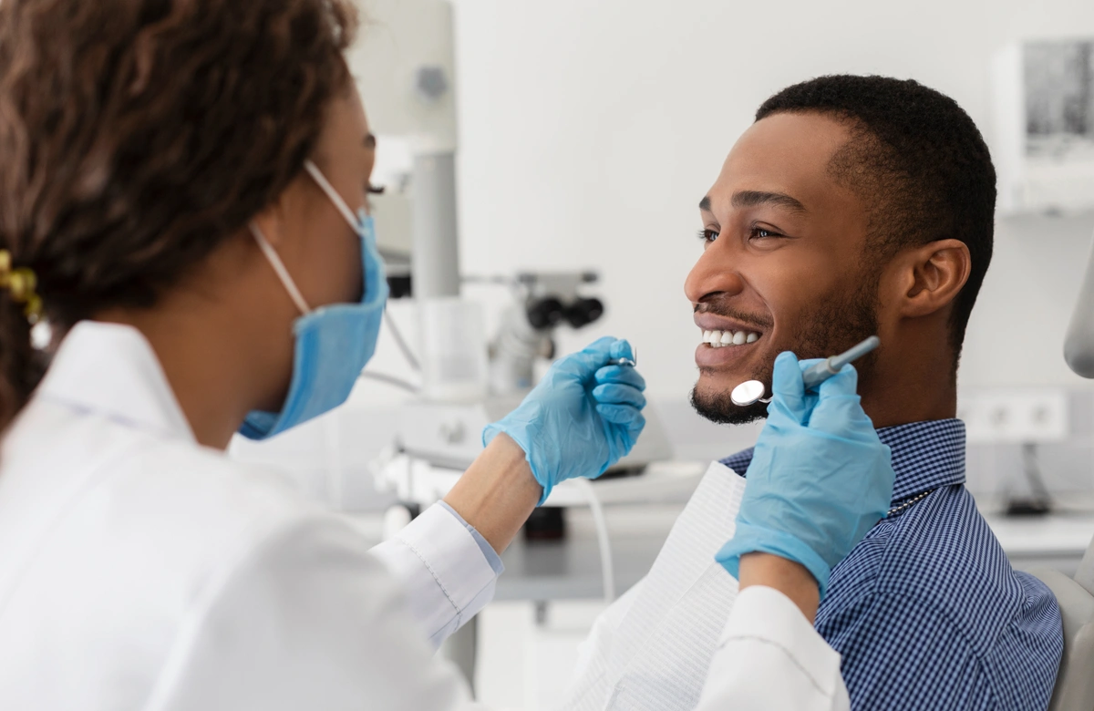 A dentist wearing a mask and gloves examines a smiling patient seated in a dental chair, checking for signs of bruxism. The dentist uses dental tools to assess the patient's teeth in a bright, modern office.