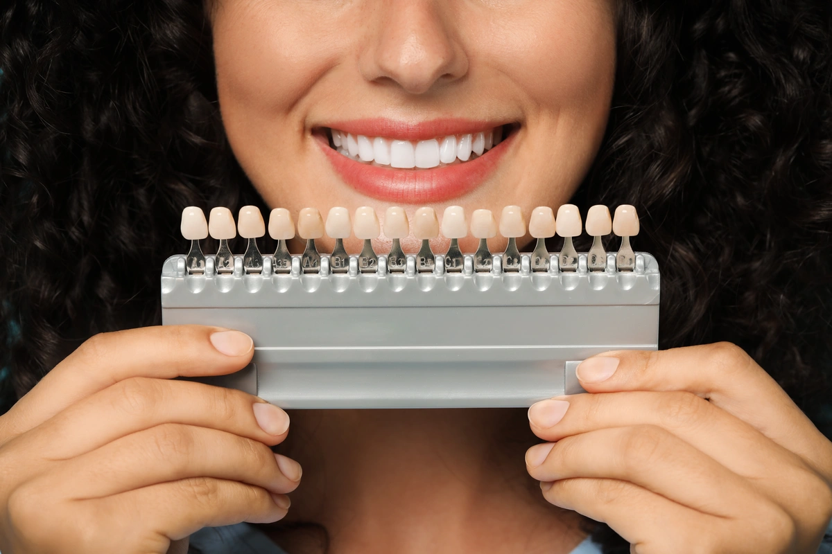 A person with curly hair smiles while holding a set of dental shade guides in front of their mouth, showcasing various tooth colors. Their teeth are bright and white, providing a comparison to the shade samples, hinting at the potential benefits and affordability of porcelain veneers with insurance.