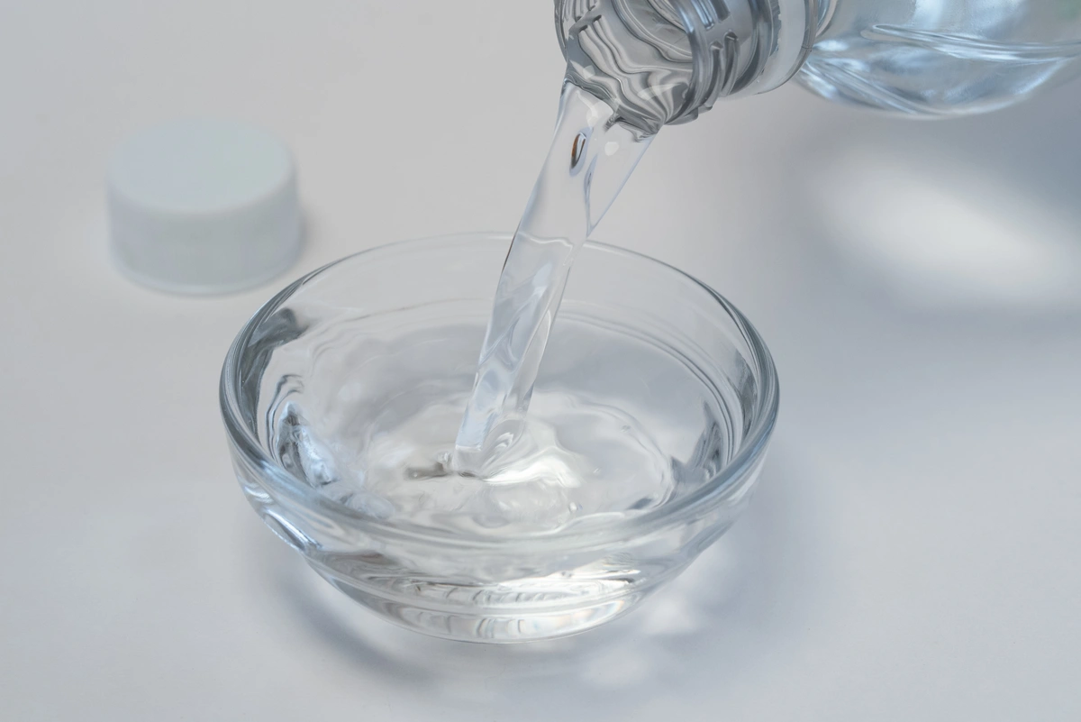 Clear water being poured from a bottle into a transparent glass bowl on a white surface, resembling the freshness you aim for when learning how to remove plaque from teeth. The bottle cap is visible nearby.