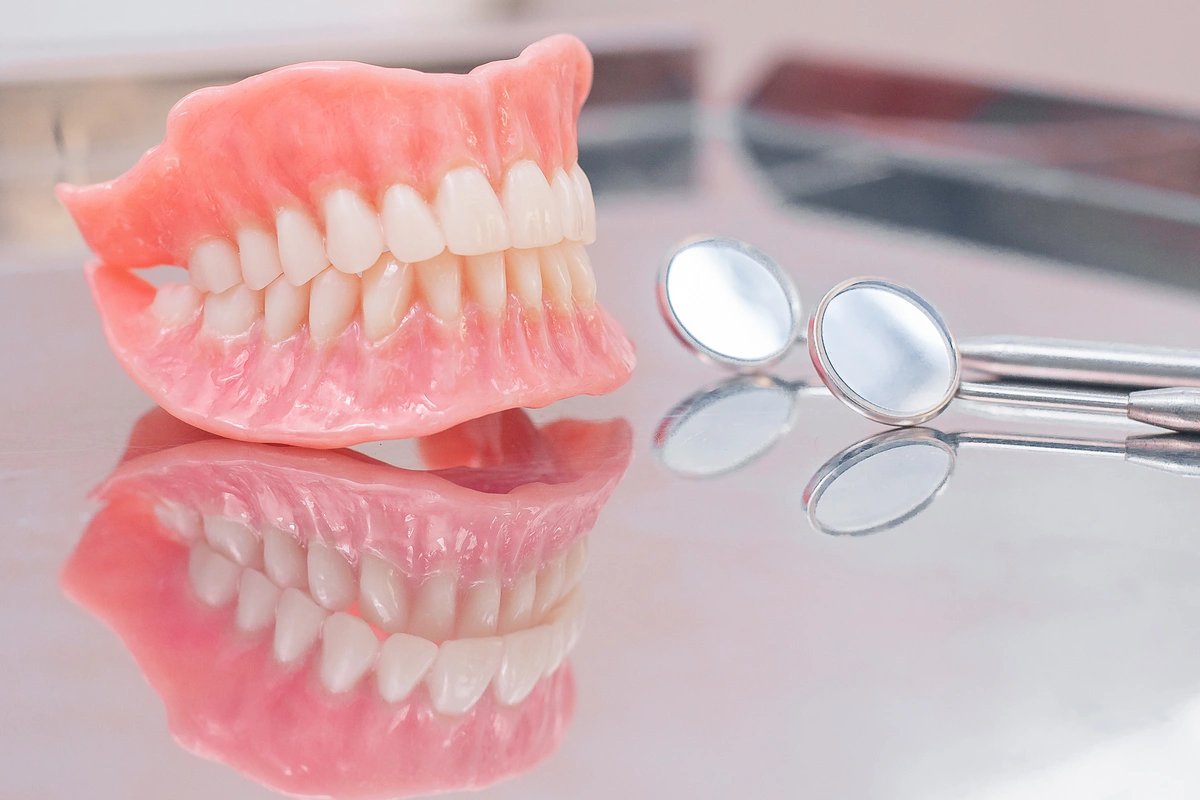A set of pink and white dentures rests on a reflective surface beside two dental mirrors. The realistic design of the teeth and gums is beautifully highlighted, leaving one to ponder not just their craftsmanship but also how much dentures might cost.