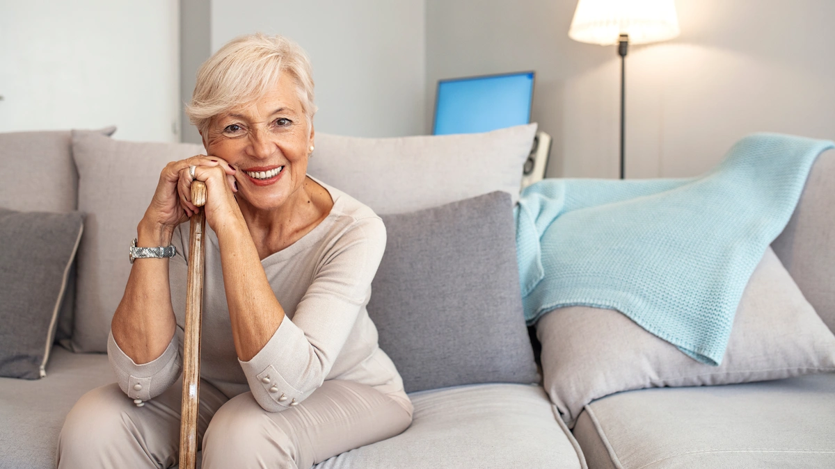 Elderly woman with short gray hair smiles, sitting on a light gray sofa. She rests her chin on a wooden cane, perhaps pondering how much dentures cost. The sofa has blue and gray cushions, and a blue blanket is draped over it. A tall lamp and a computer monitor are in the background.