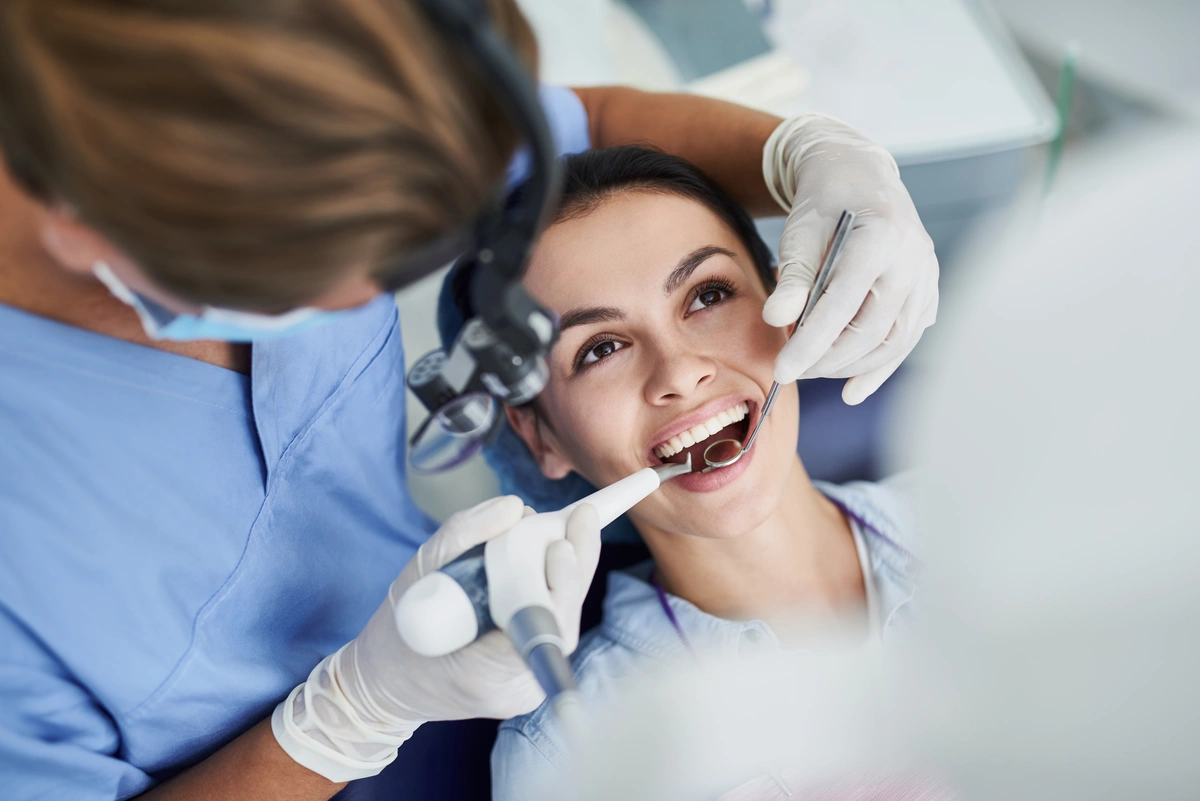 A dentist wearing blue scrubs and gloves examines a smiling woman's teeth using dental tools. The woman is lying back in a chair, and the dentist is focused on her open mouth.