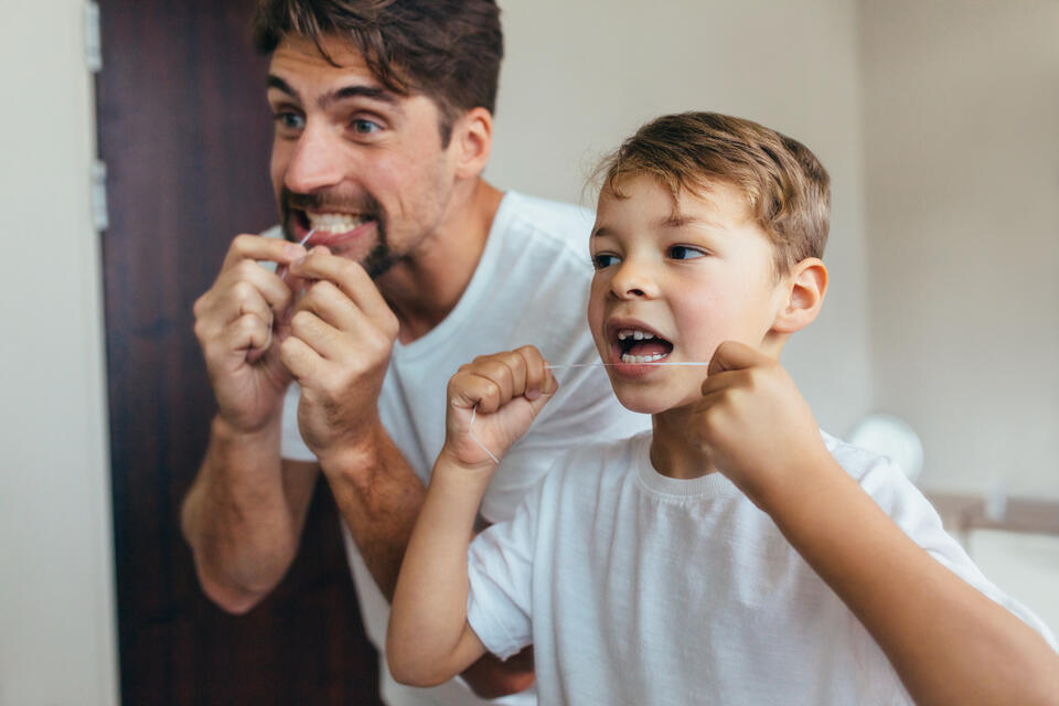 A man and a young boy stand side by side in a bathroom, smiling and flossing their teeth in front of a mirror. Both are wearing white shirts and appear to be enjoying the activity.