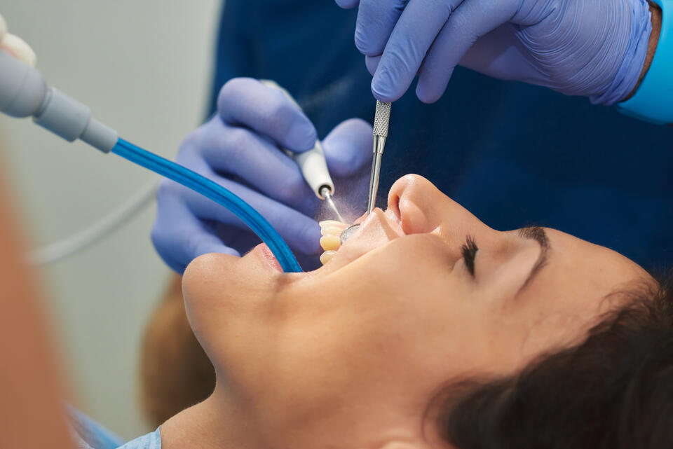 A dental patient lying back while a dentist in blue gloves works on their teeth using dental tools, including a scaler and suction. The patient’s mouth is open, and the focus is on the dental procedure.