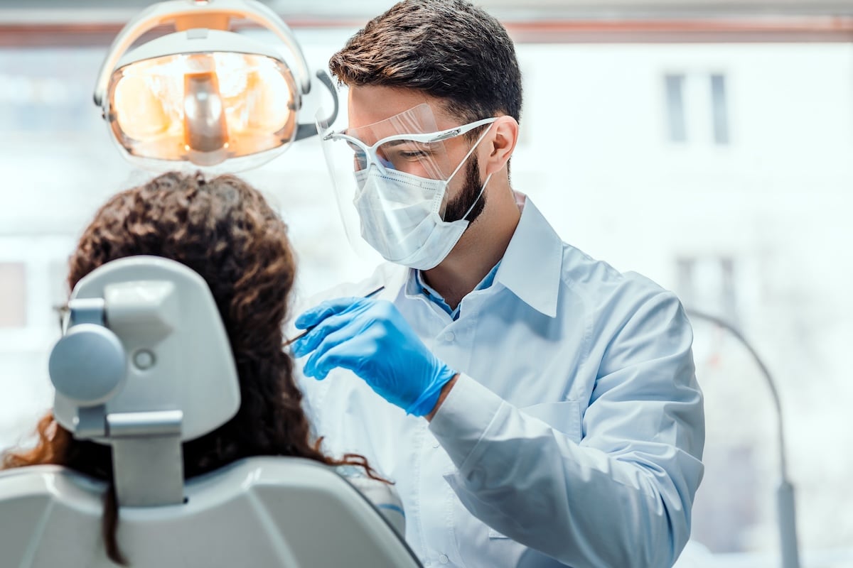 A dentist wearing a mask, safety glasses, and gloves examines a patient seated in a dental chair under an overhead light in a dental clinic.