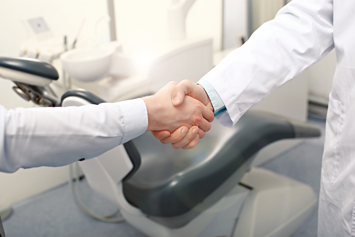 Two individuals in white lab coats shake hands in a dental office, with a dental chair in the background. The setting is bright and professional.