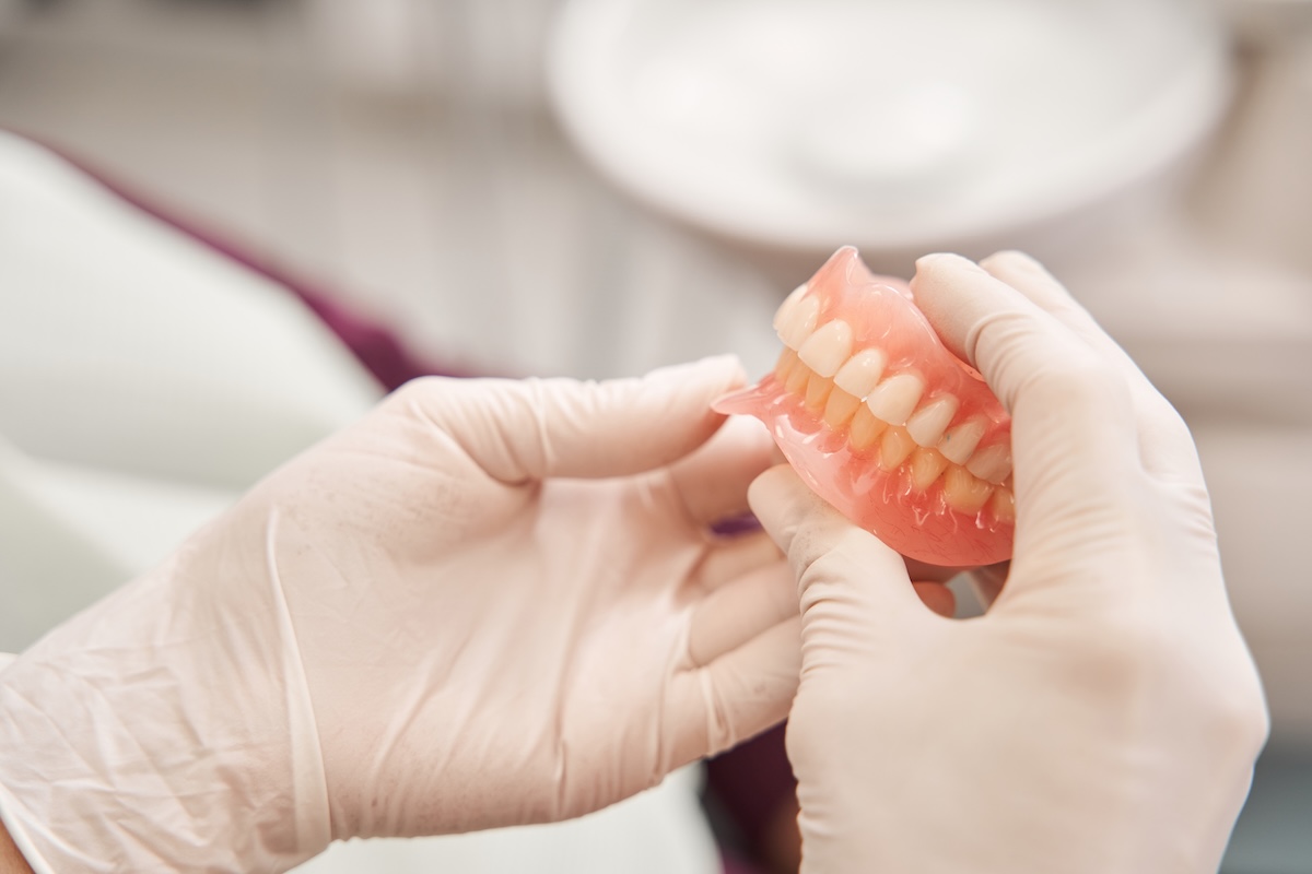 A person wearing white medical gloves holds a set of artificial dentures, showing both upper and lower teeth, in a dental or medical setting.