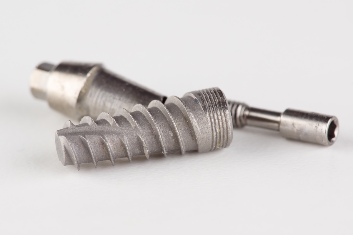 Two metallic dental implants with screw-like threads are lying on a white surface. One implant is positioned horizontally in the foreground, while the other is slightly behind and angled.