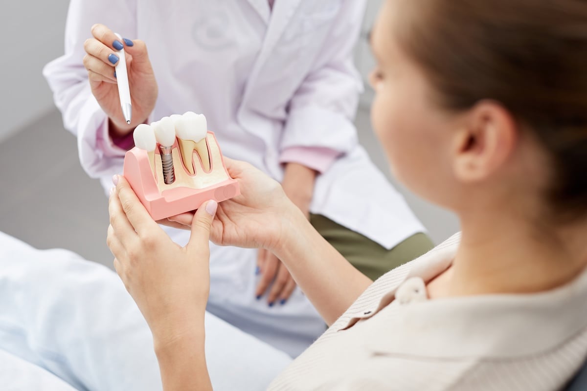 A person holds a dental model showing a dental implant while a dentist, partially visible in a lab coat, explains the model using a dental tool.
