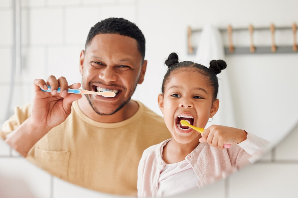 A father and daughter are smiling while brushing their teeth together in front of a bathroom mirror