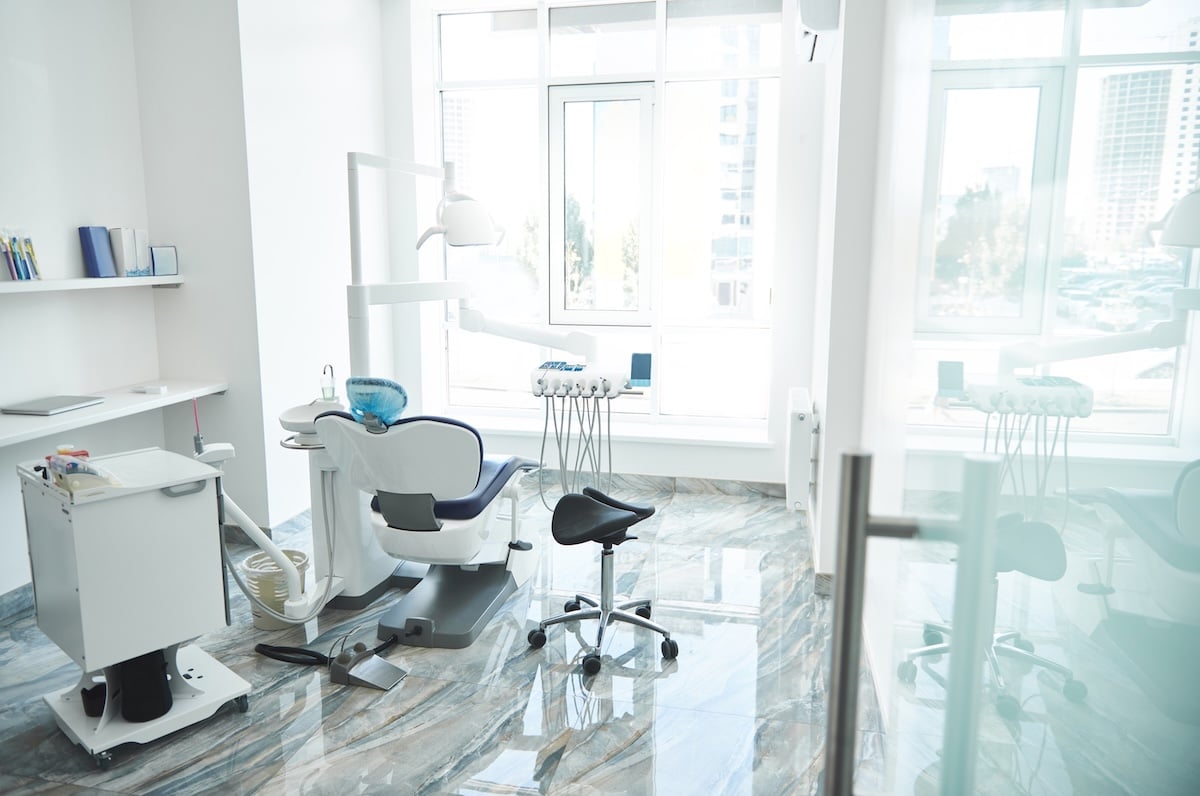A bright, modern dental clinic room with a dental chair, equipment, stool, and large windows letting in natural light, reflecting on the shiny floor.
