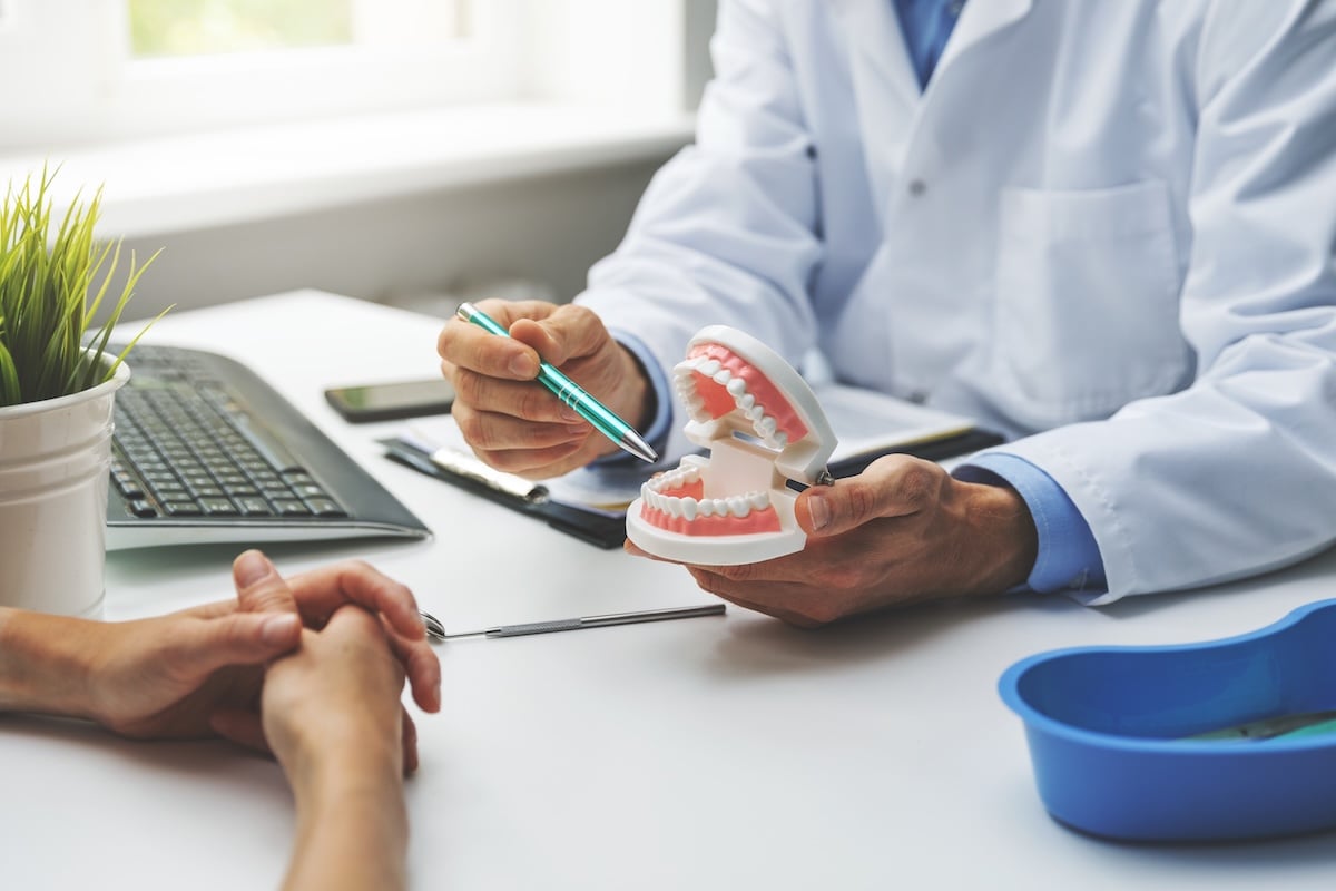 A dentist in a white coat uses a pen to point at a dental model while explaining dental care to a patient seated across a desk