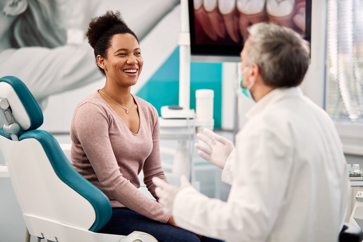 A woman sits in a dental chair smiling and talking with a dentist who is wearing a white coat, gloves, and a face mask, in a brightly lit dental office