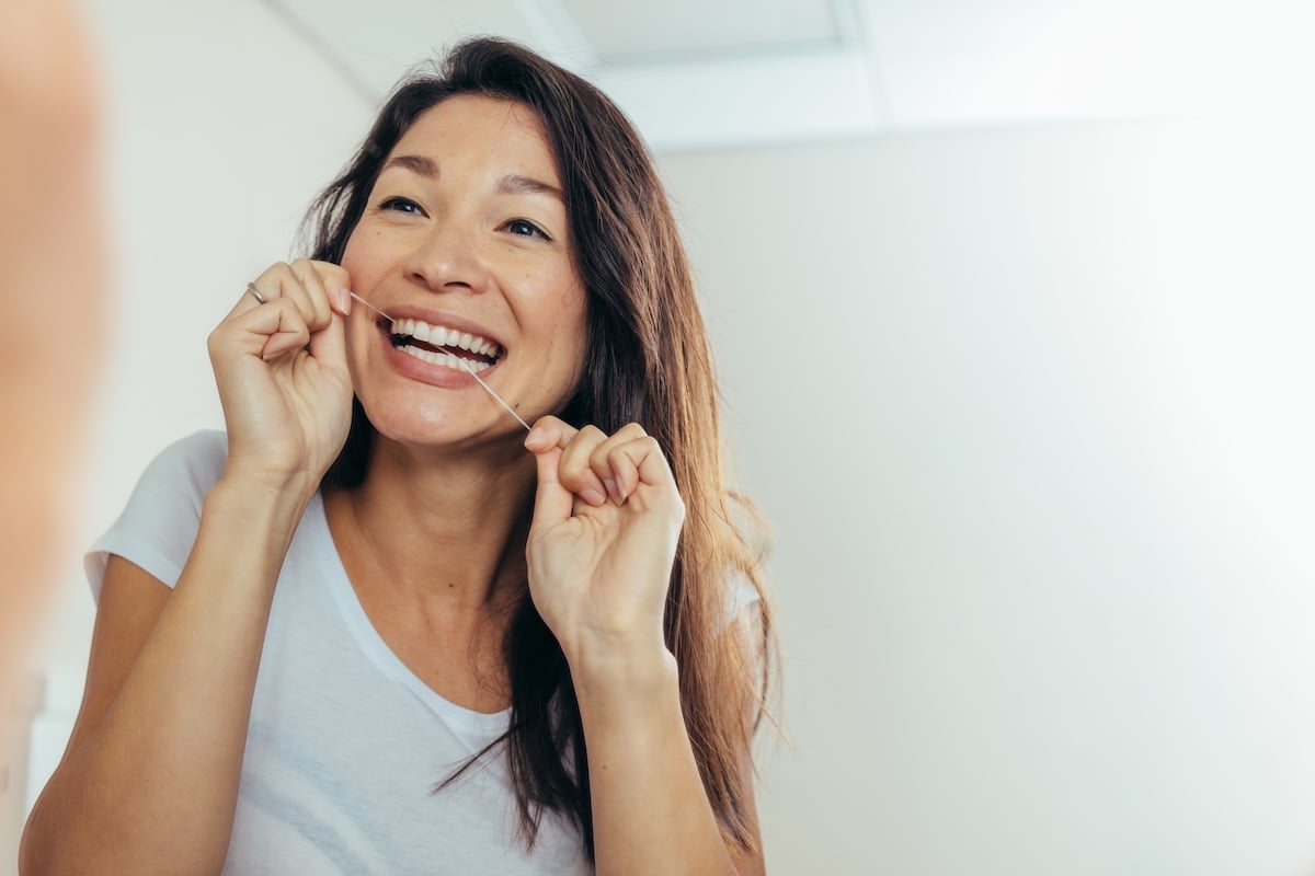 A woman with long brown hair and a white t-shirt is smiling while flossing her teeth in front of a bathroom mirror.
