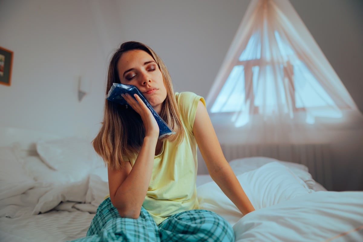 A woman in pajamas sits on a bed holding an ice pack to her cheek, eyes closed, appearing to be in pain or discomfort