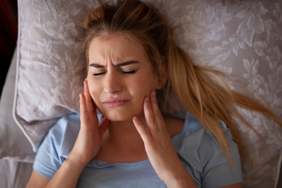 A woman lies on a pillow with a pained expression, eyes closed, and hands pressing both sides of her jaw