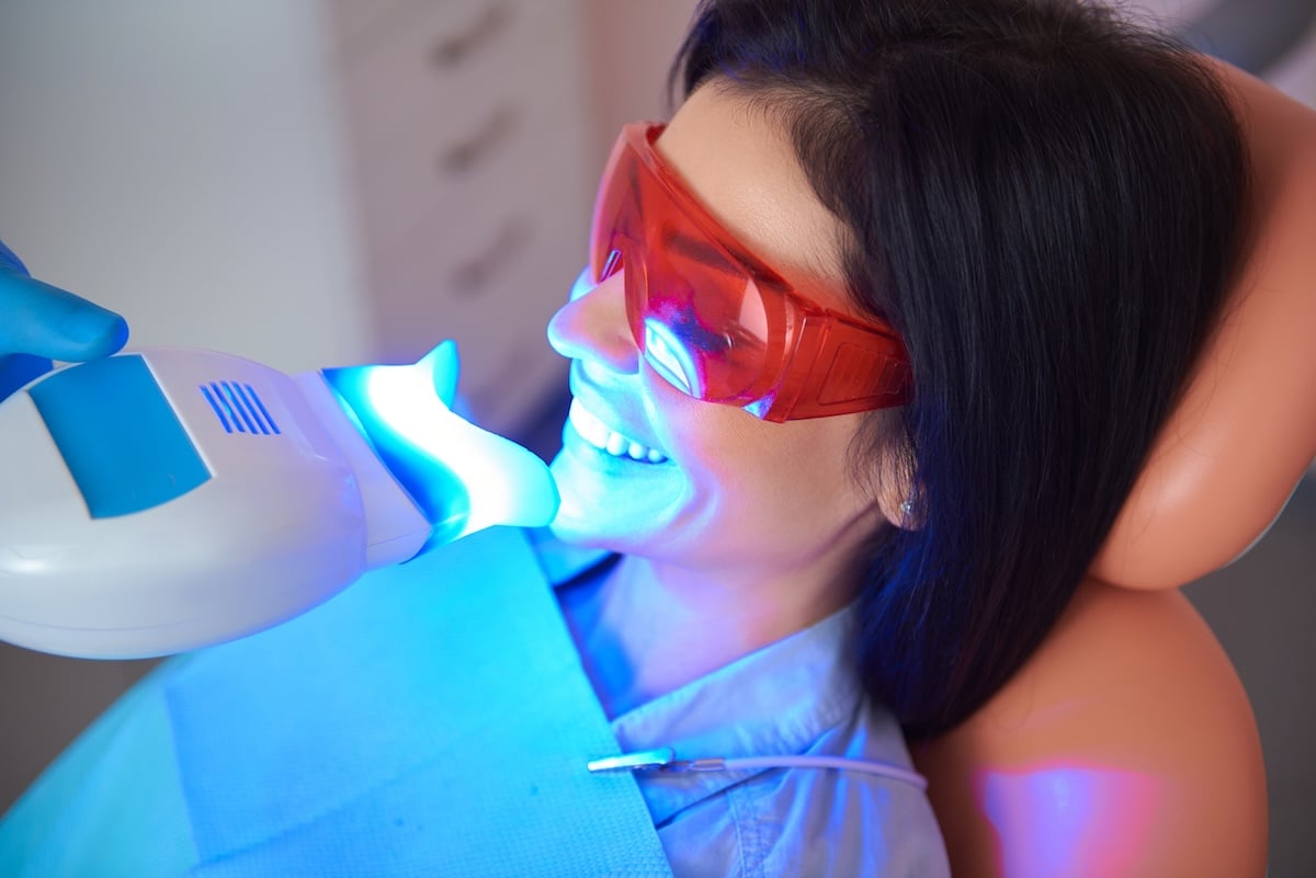 A woman wearing red protective glasses receives a teeth whitening treatment at a dental clinic, with a blue light device held near her mouth.