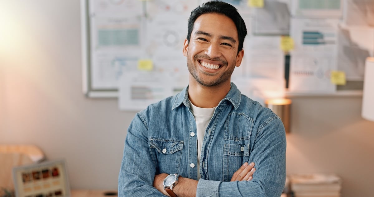 A man with dark hair and a beard, wearing a denim shirt and white t-shirt, stands smiling with arms crossed in an office setting
