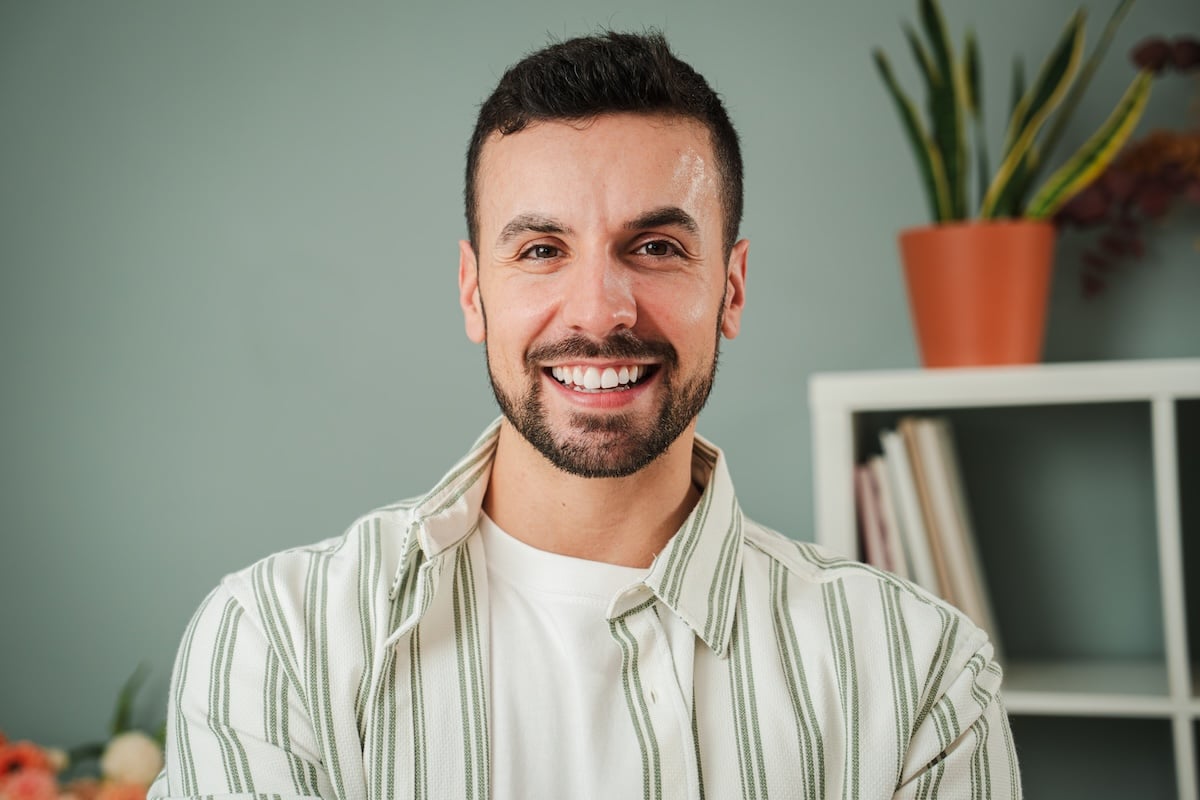 A smiling man with short dark hair and a beard stands indoors in front of a green wall, wearing a white shirt with green stripes.
