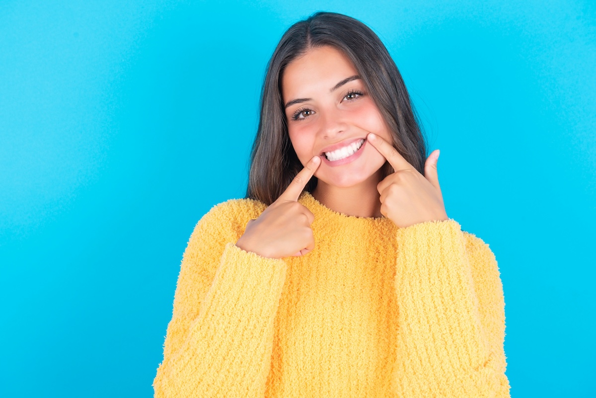 A young woman wearing a yellow sweater stands against a bright blue background, smiling widely and pointing to her teeth with both index fingers.