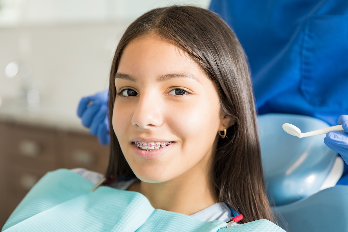 A teenage girl with long dark hair and braces smiles while sitting in a dental chair