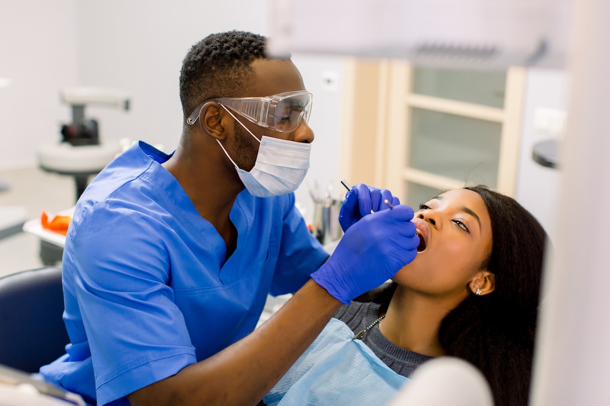 A dentist wearing safety goggles, a mask, and gloves examines a patient’s mouth with dental tools while the patient reclines in the dental chair.