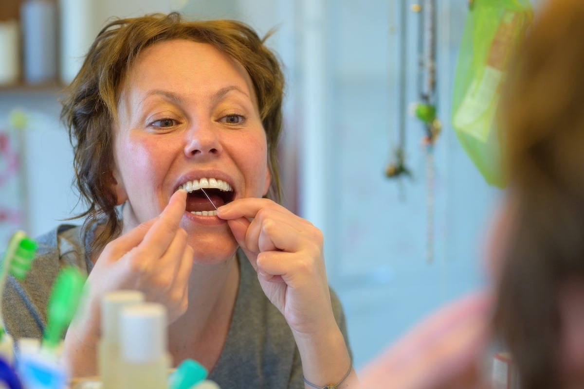 Woman flossing her teeth in a bathroom mirror using dental floss