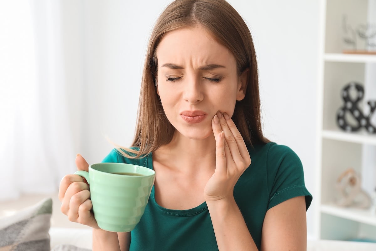 A woman in a green shirt holds a green mug and touches her cheek with a pained expression