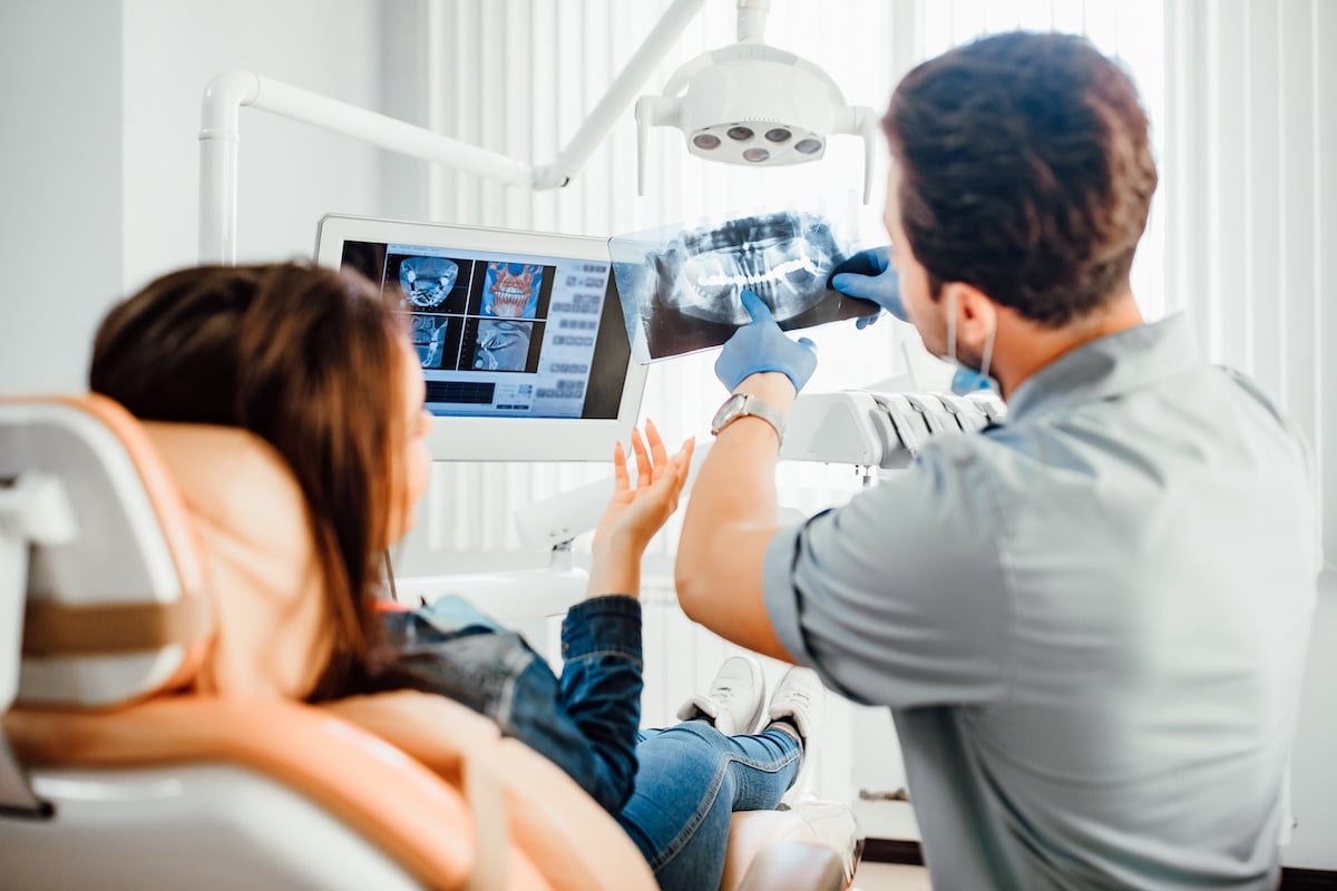 A dentist shows a dental X-ray to a patient sitting in a dental chair as they discuss urgent dental care needs, reviewing the image displayed on a computer screen in a brightly lit dental office.