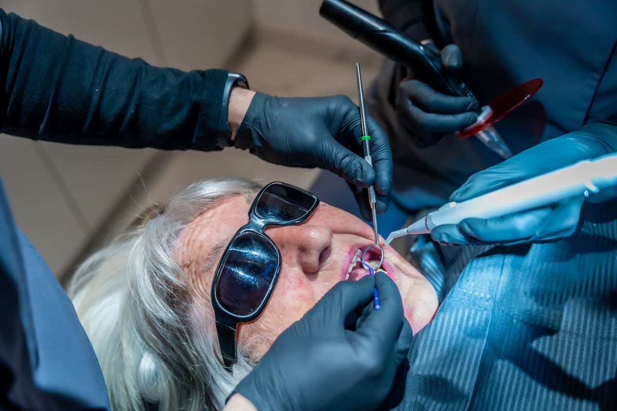 Dental professionals using specialized tools are performing a teeth whitening procedure on a senior woman patient wearing protective eyewear