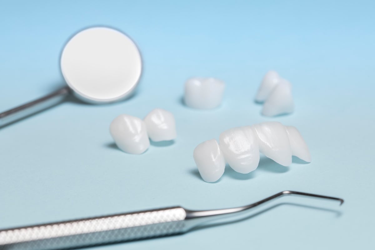 Dental tools, including a mirror and pick, lie on a light blue surface next to several white dental crowns or veneers, representing dental restoration materials.