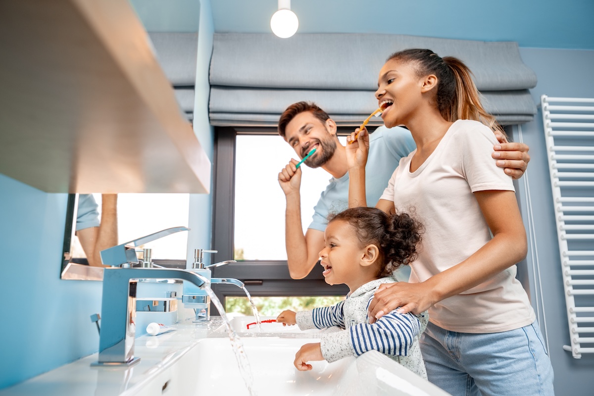 A family of three stand together in a brightly lit bathroom, smiling and brushing their teeth