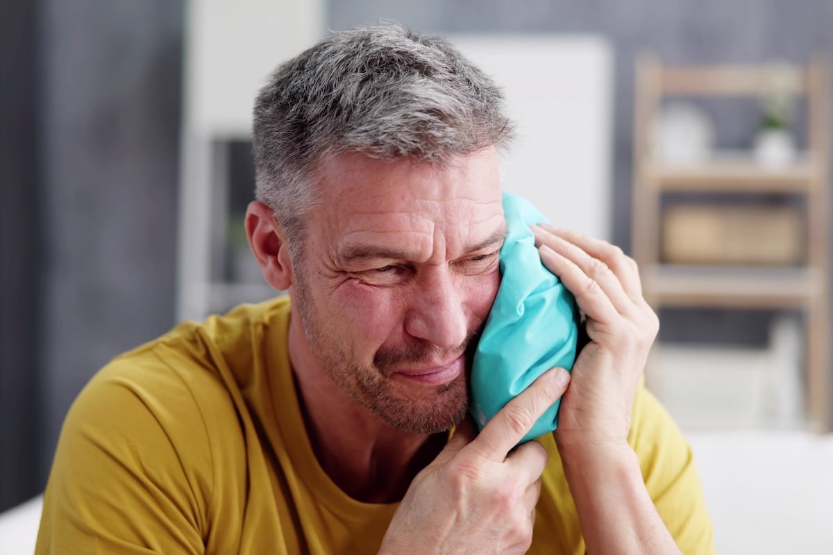 A middle-aged man in a yellow shirt holds a blue ice pack to his cheek
