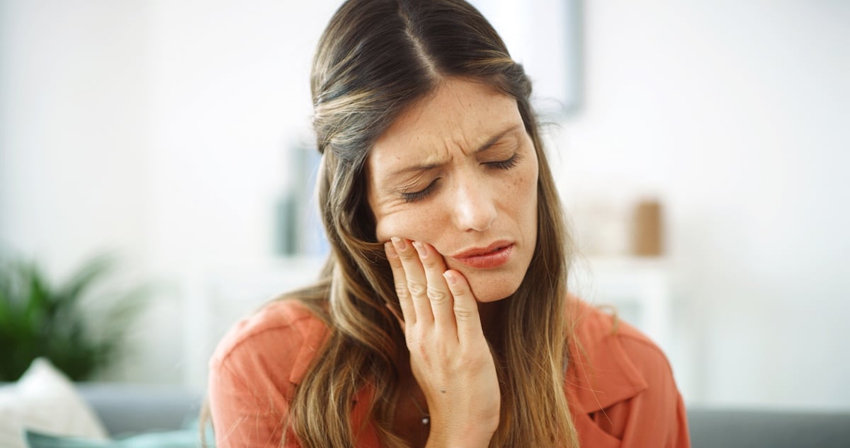 A woman with long brown hair touches her cheek and closes her eyes, appearing to be in pain, possibly due to a toothache.