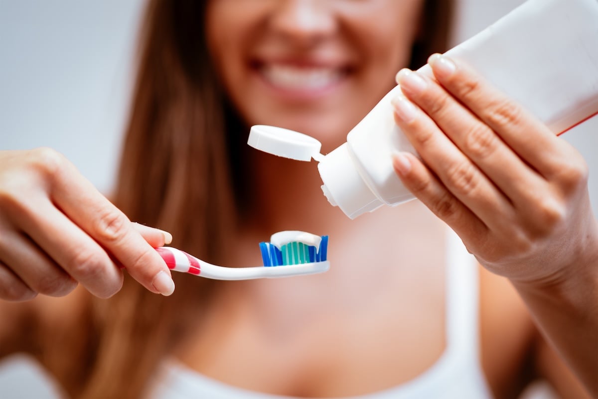 A smiling woman holds a toothbrush and squeezes toothpaste onto it, preparing to brush her teeth. 