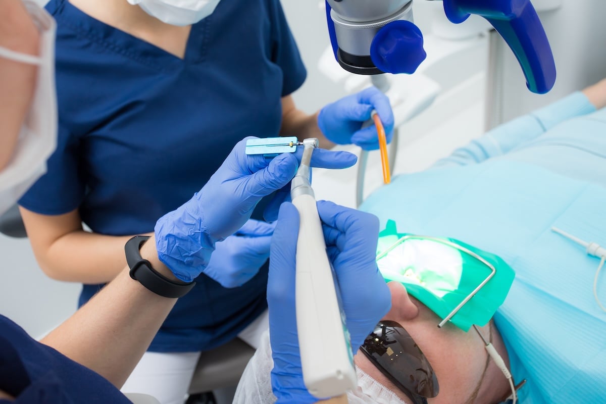 A patient undergoing a dental procedure lies on a chair wearing protective glasses. Two dental professionals in blue scrubs and gloves use instruments and a microscope, with the patient’s mouth covered by a green dental dam.