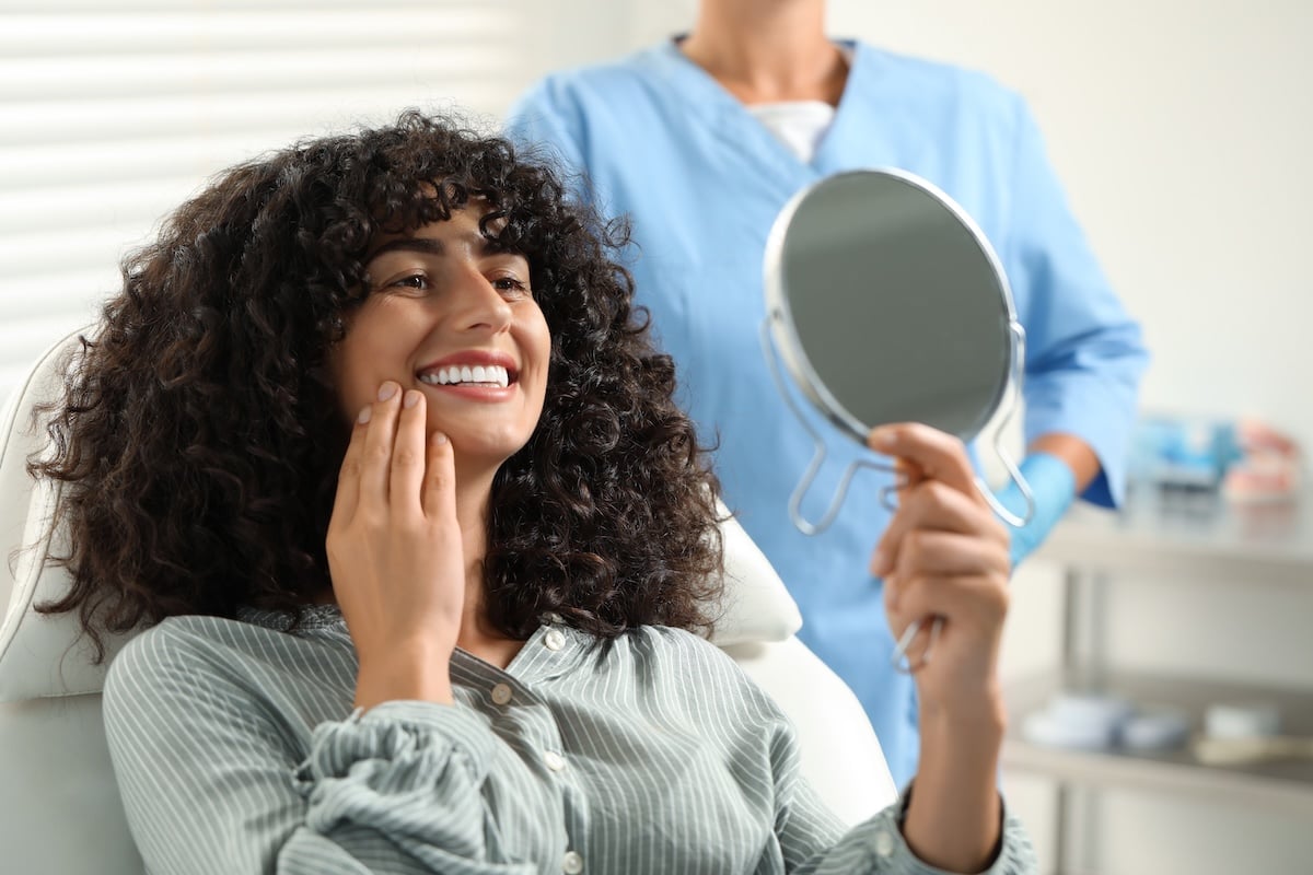 A woman with curly hair smiles while looking at her reflection in a handheld mirror, sitting in a dental chair.