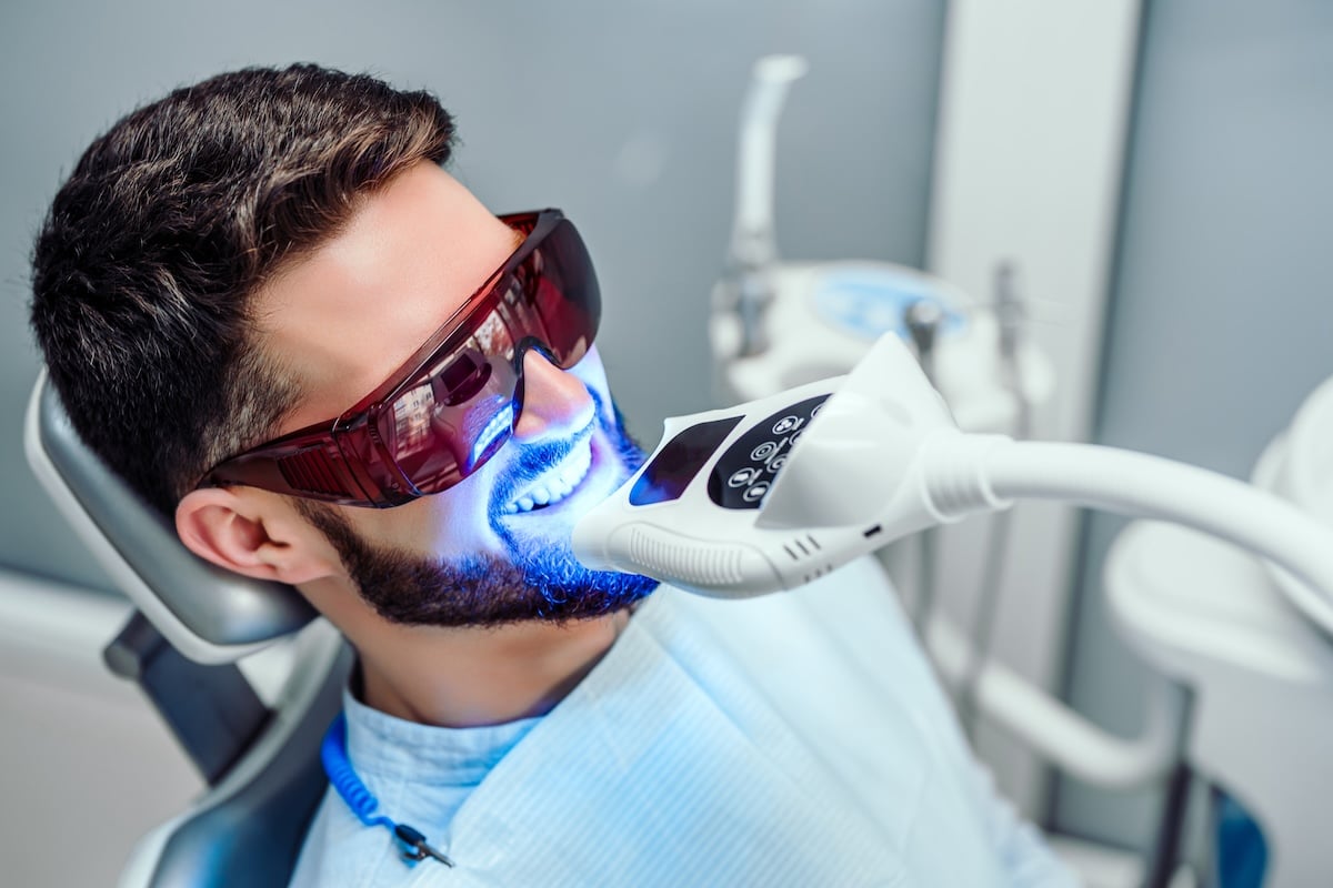 A man wearing protective red glasses sits in a dental chair while undergoing a teeth whitening procedure